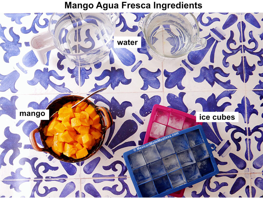Overhead view of mango agua fresca ingredients on a blue and white tile: a bowl of diced mango, a pitcher of water, and two ice cube trays with ice. Each ingredient is labeled.