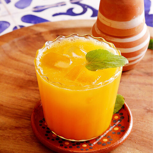 A glass of bright mango agua fresca with ice and a fresh mint leaf garnish, placed on a round coaster on a wooden surface. A striped clay cup is in the background, and blue patterned tiles are partially visible.
