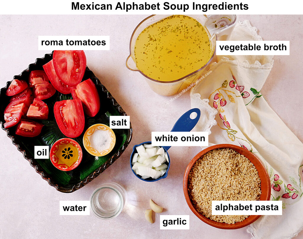 Ingredients for Mexican alphabet soup are arranged on a light surface: roma tomatoes, a cup of vegetable broth, salt, oil, chopped white onion, a bowl of alphabet pasta, water, and garlic cloves.