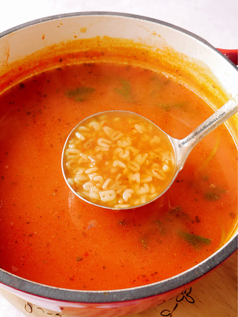 A pot of bright red Mexica alphabet soup with pasta letters floating in a tomato-rich broth, a ladle lifting a scoop of the flavorful soup above the surface.