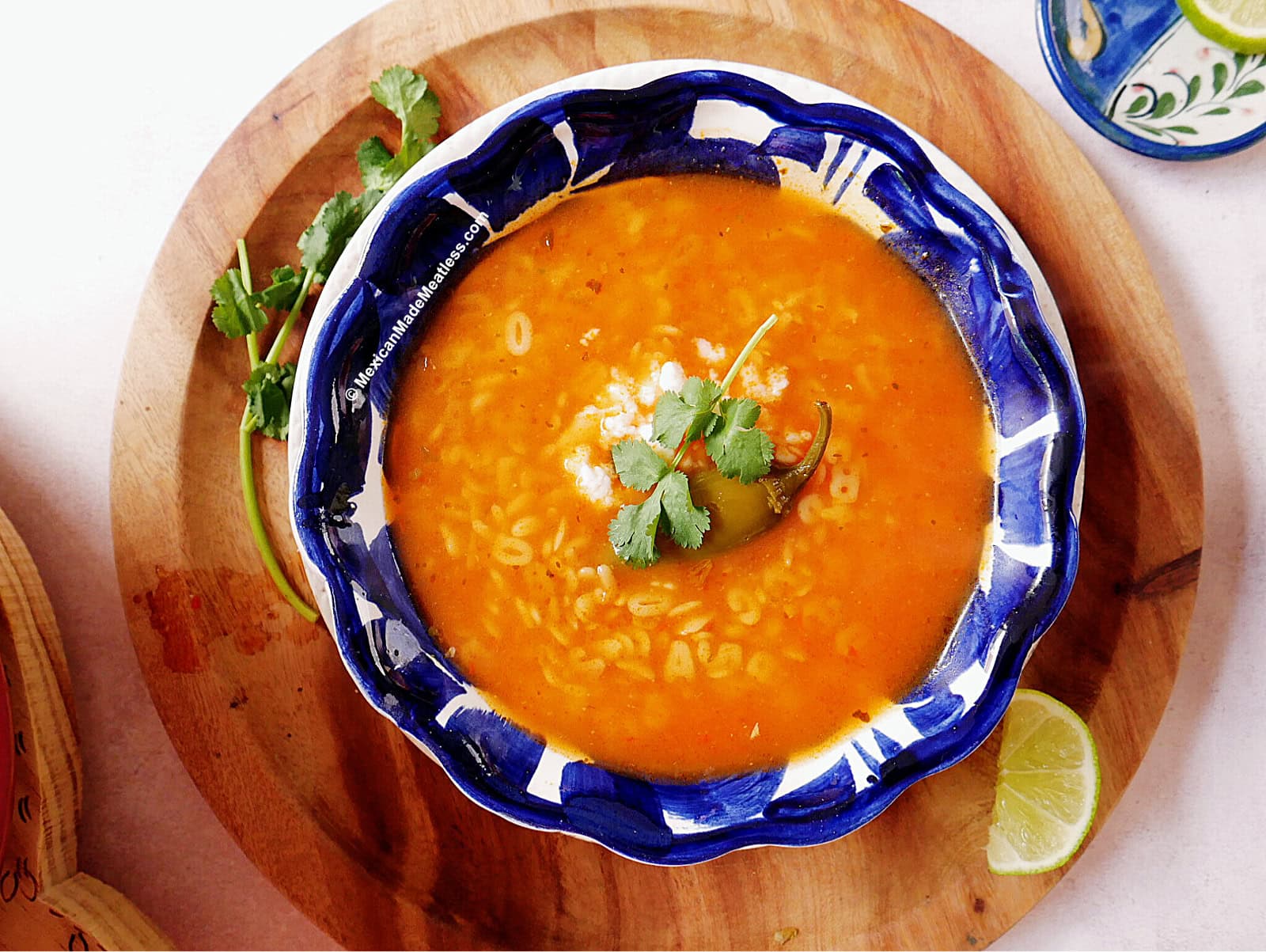 A blue and white bowl of Mexican alphabet soup filled with tomato broth, pasta, and a pickled jalapeno pepper, garnished with cilantro and crumbled cheese. The bowl sits on a wooden board with a sprig of cilantro and a lime wedge nearby.