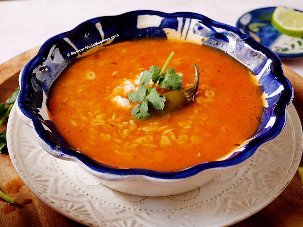 A bowl of Mexican sopa de letras with a tomato broth, garnished with cilantro, cheese, and a pickled jalapeno pepper, served in a blue and white bowl on a decorative plate.