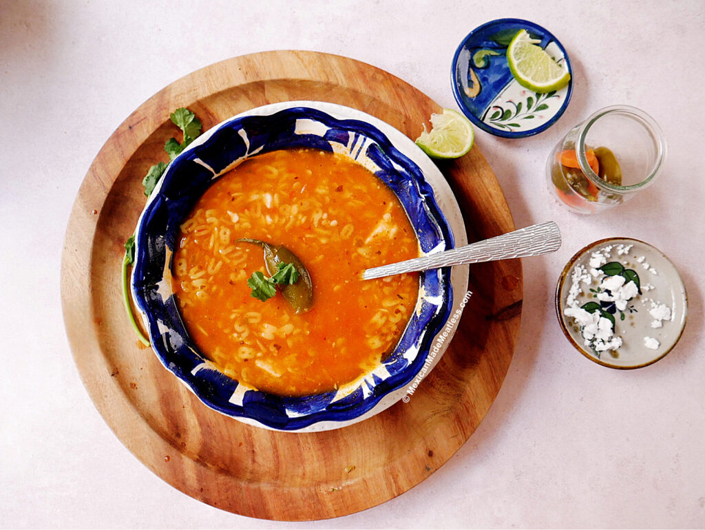 A blue and white bowl filled with Mexican ABC Soup sits on a wooden board, garnished with cilantro. Nearby are lime wedges on a small plate, a glass jar with jalapenos, and a small dish topped with crumbled cheese.