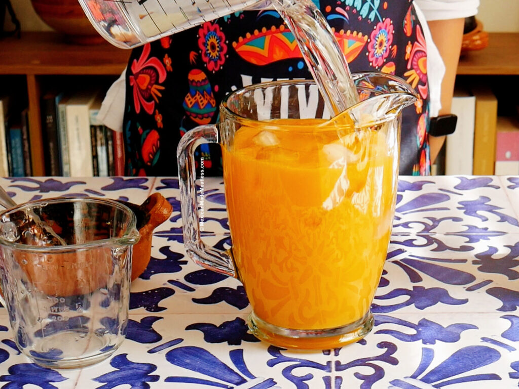 Nancy Lopez pours water into a glass pitcher filled with agua fresca de mango on a blue and white patterned table.