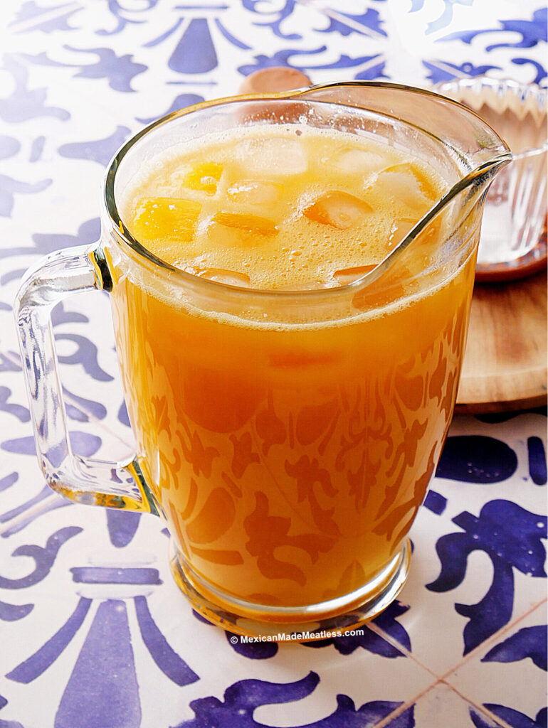 A glass pitcher filled with mango agua fresca and ice cubes sits on a blue and white patterned table. A small part of another glass and a wooden tray are visible in the background.