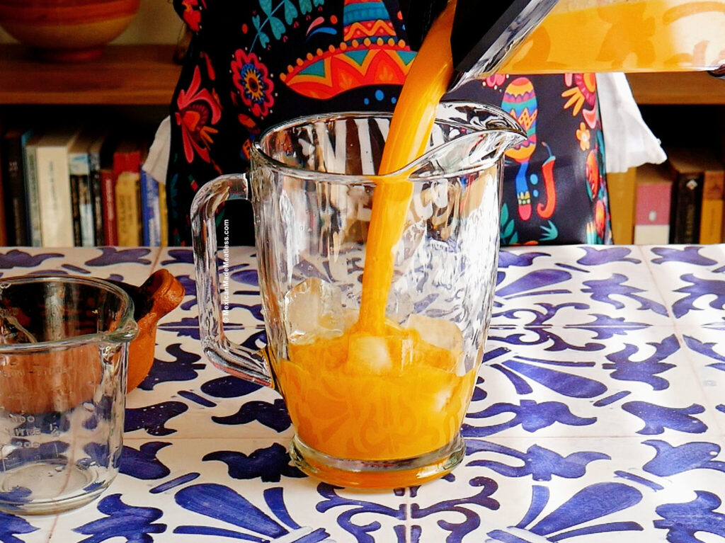 Mango agua fresca being poured from a blender into a clear glass pitcher with ice cubes on a blue and white patterned table, with books and a colorful patterned apron in the background.