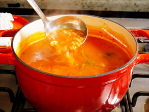 A ladle scoops up Mexican alphabet soup in a rich tomato broth, from a red pot on the stovetop. Steam rises from the hot, comforting dish.