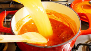 A stream of yellow vegetable broth is being poured into a red pot of Mexican ABC soup on the stove, with a wooden spoon resting inside the pot.
