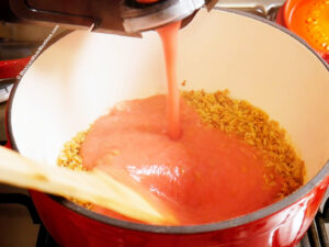 A close-up of tomato sauce being poured over toasted alphabet pasta in a pot on the stove, with a wooden spoon stirring the mixture to make soup.