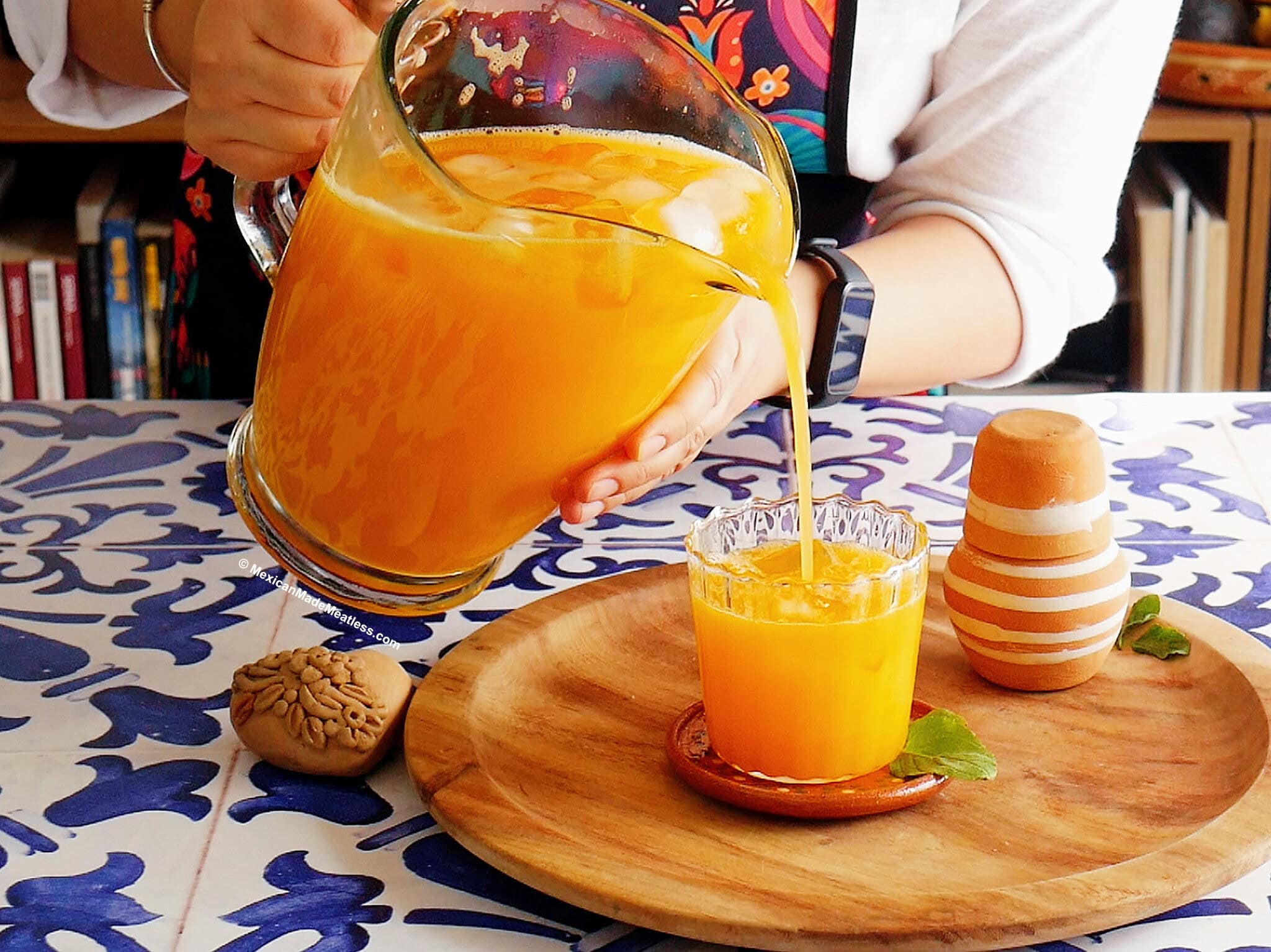 Nancy Lopez pours mango agua fresca from a glass pitcher into a glass on a wooden tray with a clay cup and mint leaves, all on a blue and white tiled table.