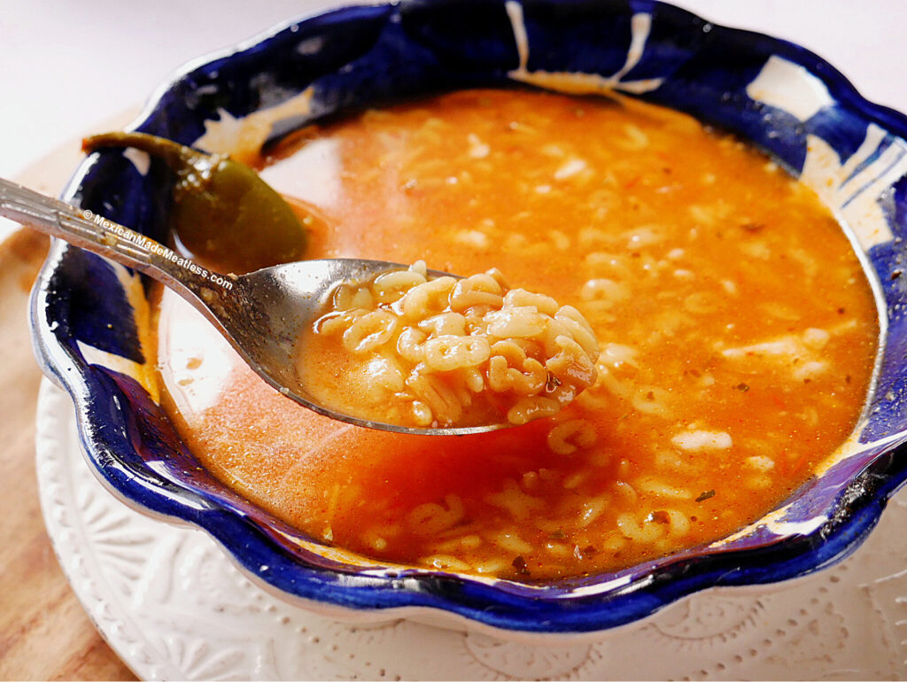 A blue ceramic bowl filled with Mexican alphabet soup sits on a white plate. A spoon lifts some of the pasta letters from the tomato-rich broth, and a whole jalapeno pepper is visible in the bowl.