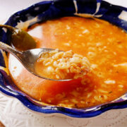 A blue ceramic bowl filled with Mexican alphabet soup sits on a white plate. A spoon lifts some of the pasta letters from the tomato-rich broth, and a whole jalapeno pepper is visible in the bowl.