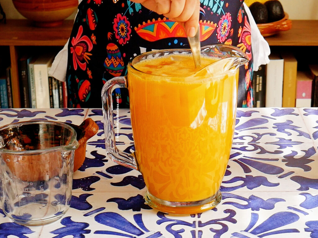 Nancy Lopez stirs a large glass pitcher of mango agua fresca on a blue and white patterned tablecloth, with colorful patterned clothing visible and a bookshelf in the background.