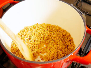 Toasted alphabet pasta being stirred with a wooden spoon in a large red pot on a stovetop.