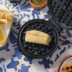 A tamale sits on a black waffle maker, surrounded by colorful dishes and food on a blue and white patterned table.