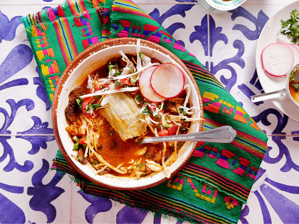 A bowl of red pozole with cabbage, radish slices, and a tamale sits invitingly atop a colorful woven cloth on a blue and white patterned surface, creating an enticing scene reminiscent of traditional tamazole soup feasts.