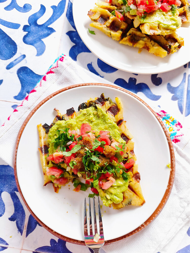 A white plate holds tamale waffle pieces topped with guacamole, diced tomatoes, herbs, and salsa. A fork rests on the plate. Another plate with food is partially visible on a colorful napkin and blue patterned table.