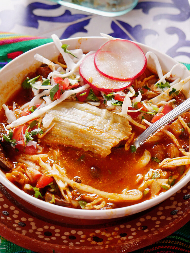 A bowl of tamazole soup made with pozole and with a tamal, sliced radishes, shredded cabbage, cilantro, and a spoon, set on a colorful table.