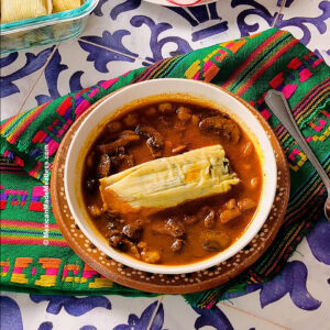 A bowl of red pozole and a tamale on top sits on a colorful woven cloth, next to a spoon on a patterned table.