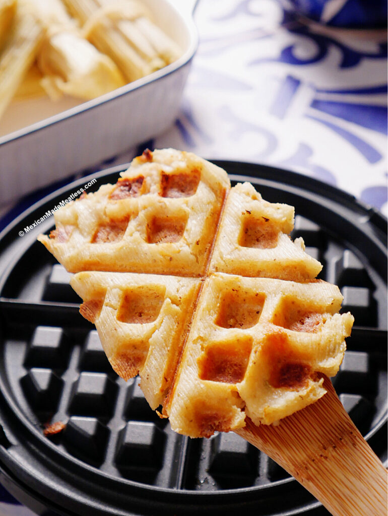 A cooked tamale waffle made with bean and cheese tamales is pressed in a waffle iron, held up on a wooden spatula, with more tamales and a waffle iron visible in the background.