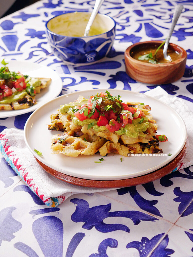 A plate a tamale waffle topped with guacamole and pico de gallo sits on a white plate. In the background are two bowls of salsa and a stack of tamales on a blue and white patterned table.
