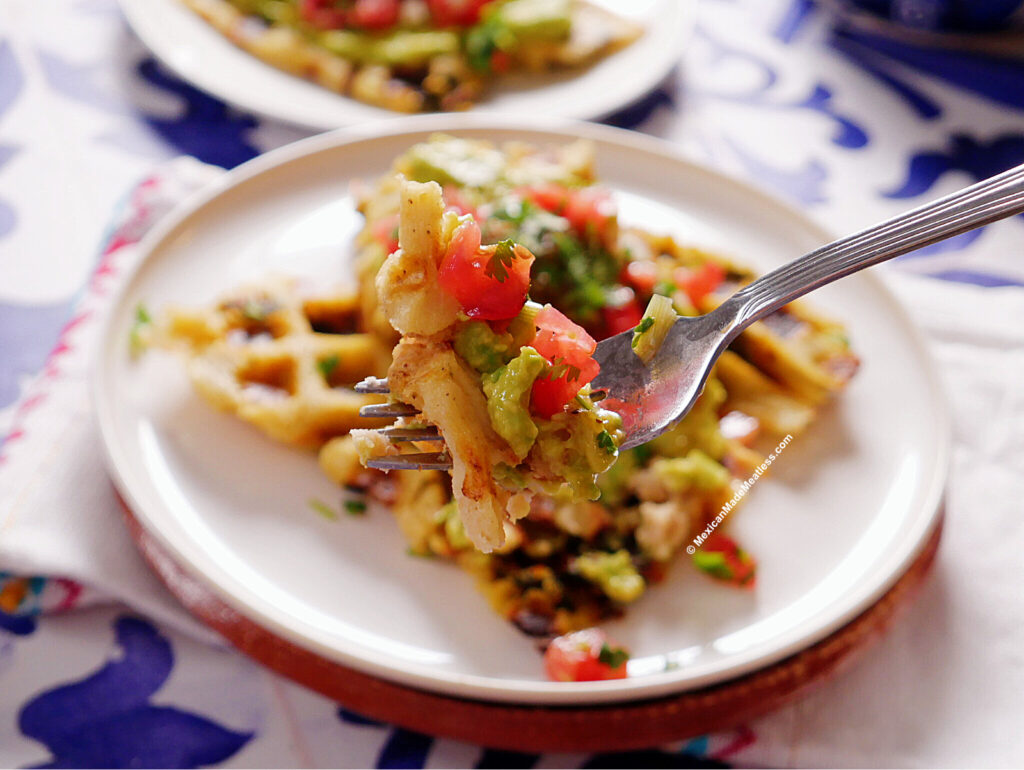 A fork holds a bite of savory tamales waffles topped with avocado and diced tomato, with more waffle and toppings on a white plate in the background.