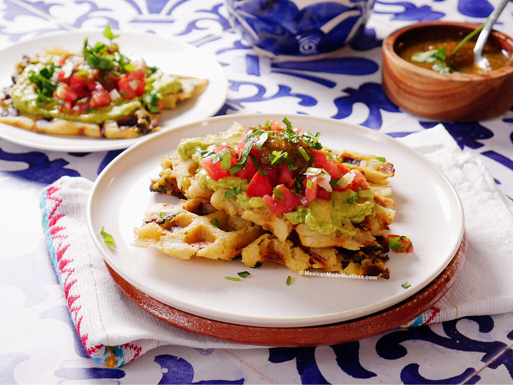 Two plates with grilled tamales waffles topped with guacamole, diced tomatoes, cilantro, and salsa, set on a blue and white tablecloth with a small bowl of green salsa nearby.