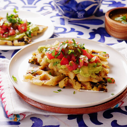 Two plates with grilled tamales waffles topped with guacamole, diced tomatoes, cilantro, and salsa, set on a blue and white tablecloth with a small bowl of green salsa nearby.