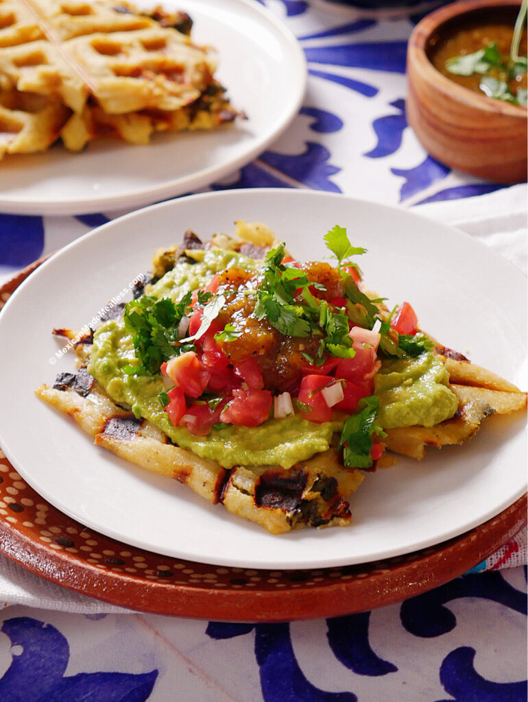 A white plate holds a tamales waffle topped with guacamole, diced tomatoes, onions, salsa, and chopped cilantro. A bowl of sauce and another serving are visible in the background on a blue-and-white patterned tablecloth.