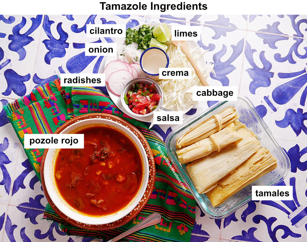 Overhead view of tamazole ingredients: pozole rojo in a bowl, tamales, limes, cabbage, crema, cilantro, onion, radishes, and salsa—all the essential tamazole ingredients—beautifully arranged on a blue patterned tablecloth.