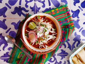 A bowl of tamale soup, is topped with sliced radishes, shredded cabbage, cilantro, and a lime wedge, set on a colorful woven cloth next to a spoon and a tamale, all arranged on a blue and white tiled surface.