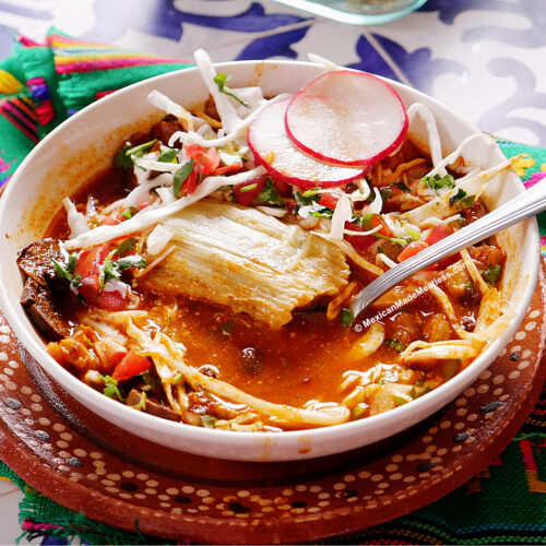 A bowl of tamazole which is a red pozole served with a tamal in the bowl, and topped with cabbage, radish slices, cilantro, with a spoon inside. The dish is set on a colorful woven cloth.