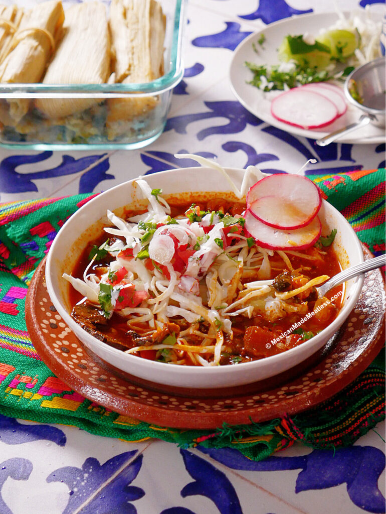 A bowl of tamazole which is pozole with a tamal inside the bowl, and topped with shredded lettuce, sliced radishes, cilantro, and onions sits on a colorful cloth. Tamales and fresh garnishes are on dishes in the background.