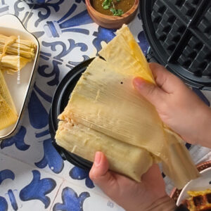 Two hands unwrap a corn husk from a tamale over a waffle iron. Nearby are more tamales, a green sauce in a bowl, and a patterned blue and white tile surface.