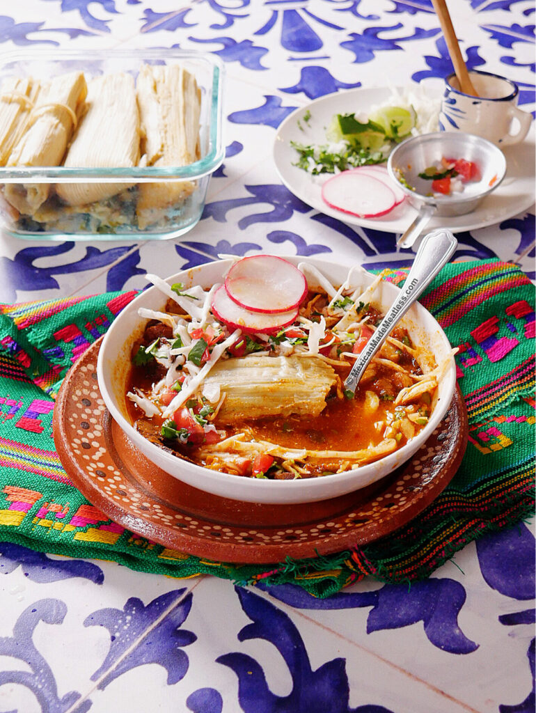 A bowl of red pozole with a tamal, topped with radish and cabbage, sits on a colorful cloth. In the background are more tamales, lime wedges, and a blue-patterned table.