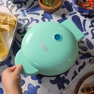 A hand holds the handle of a closed mint green Dash waffle maker on a blue and white tile table, surrounded by tamales, salsa, and other food dishes—making waffle tamales.