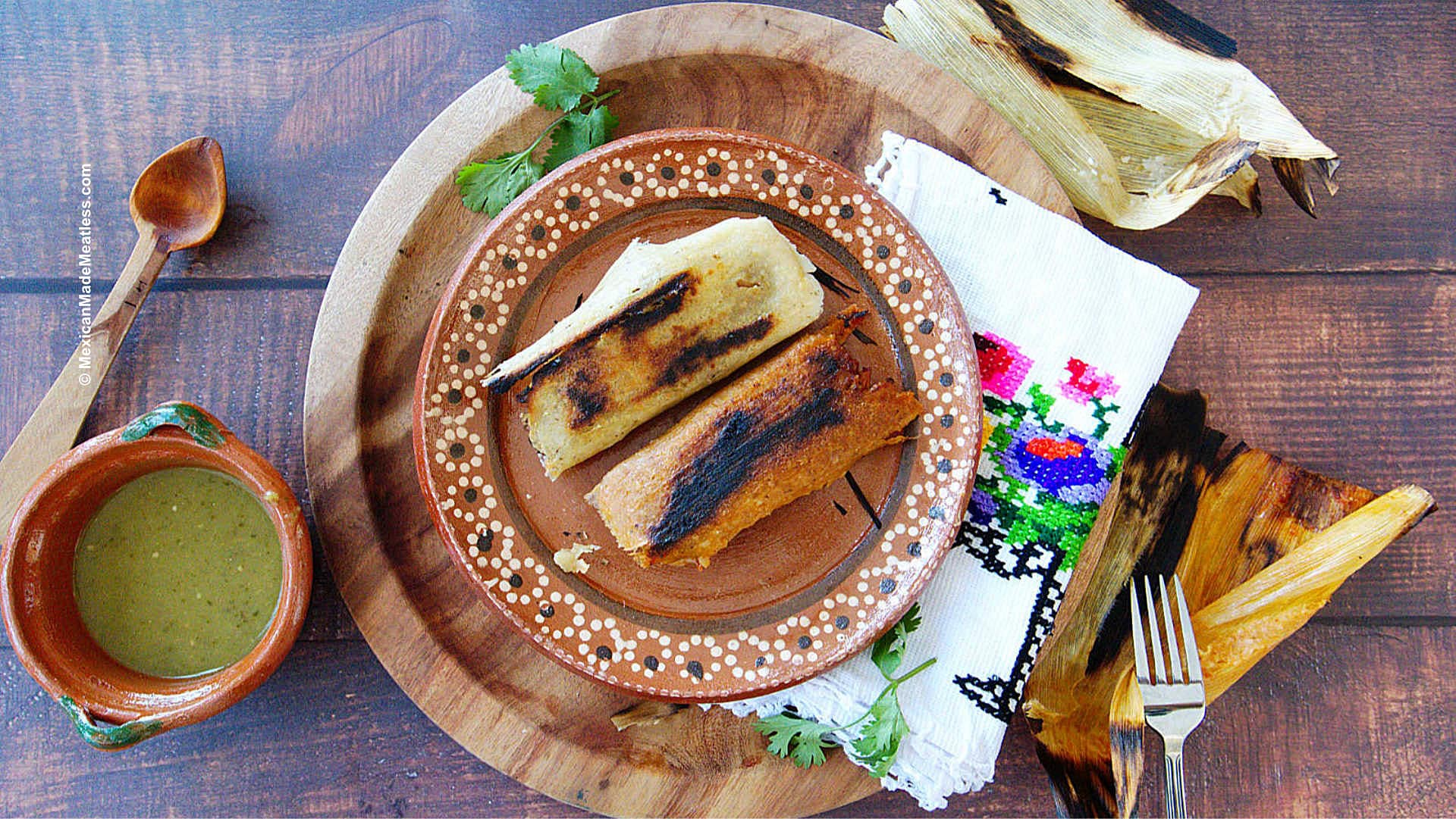 A plate with two tamales reheated with crispy texture on a wooden board, cilantro garnish, a fork, a colorful embroidered napkin, and a small bowl of green salsa on a rustic table.