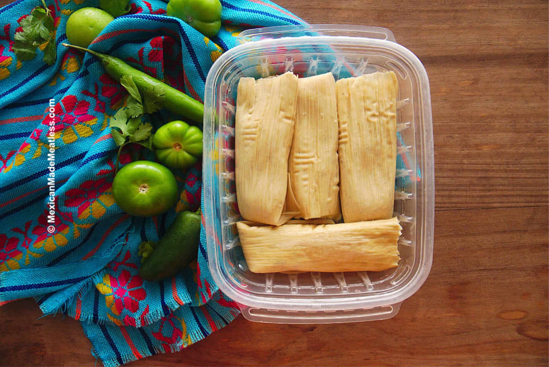A clear plastic container with five tamales sits on a wooden table next to green tomatillos, jalapeños, cilantro, and a colorful blue towel with red and pink floral patterns.